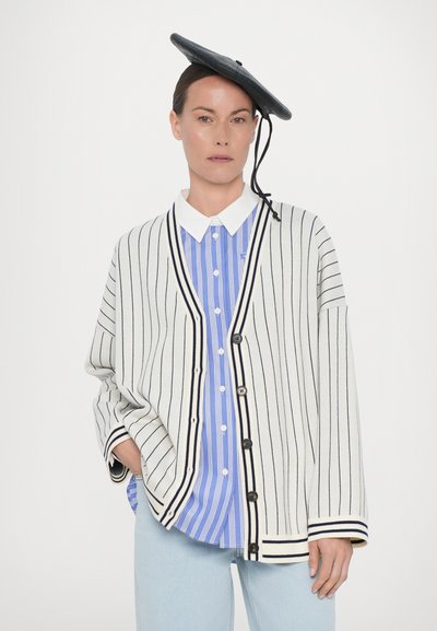 Cardigan crème rayé avec bord noir, superposé à une chemise rayée bleu et blanc. Un béret noir ajoute du contraste et un détail unique.