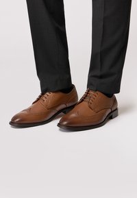 Brown leather dress shoes featuring wingtip detailing, lace-up design, and a smooth finish, paired with grey tailored trousers.