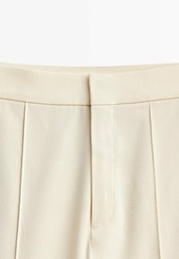 Pantaloni color crema realizzati in un tessuto liscio, con frontale plissettato e un delicato dettaglio cucito lungo la vita.
