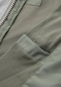 Grøn stof med en glat overflade og en meshlomme. Synlige lynlåsdetailler på den ene side, som fremviser et lagdelt design.