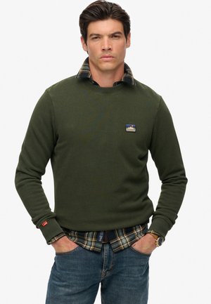 Man in een groene sweatshirt over een geruit shirt, met zijn handen in de zakken van zijn jeans, staand tegen een eenvoudige witte achtergrond.