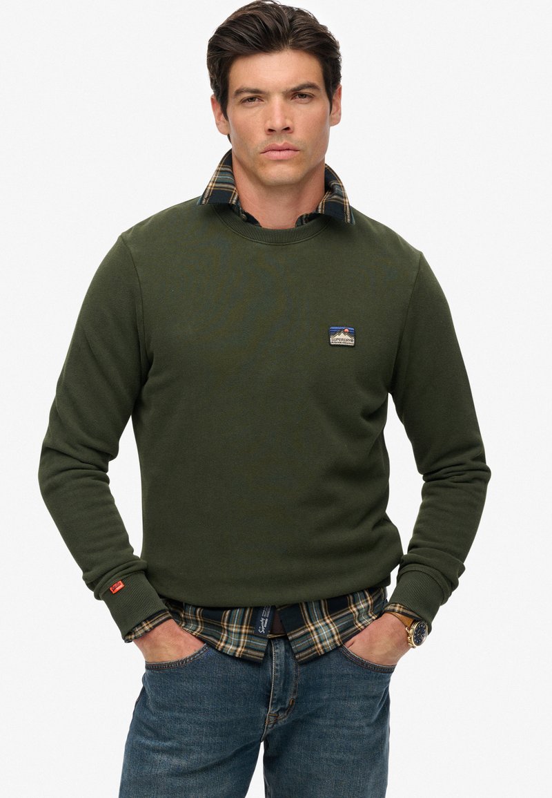 Man in een groene sweatshirt over een geruit shirt, met zijn handen in de zakken van zijn jeans, staand tegen een eenvoudige witte achtergrond.