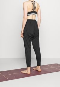 Svarta, lätta joggers med elastisk midja, tillsammans med en svart sport-bh med korsande remmar och ett grått band, på en yogamatta.