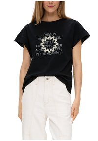 Schwarzes Baumwoll-T-Shirt mit kurzen, aufgerollten Ärmeln, das weißen Text und Vogelgrafiken in einem kreisförmigen Design zeigt. Trägt dazu helle Hosen.