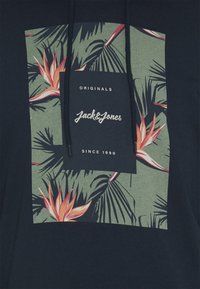 Sudadera oscura con estampado de hojas y flores tropicales en verde y naranja, con el texto "ORIGINALS Jack & Jones DESDE 1990" en el centro.