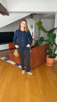 Νεαρή γυναίκα με σκουρό μπλε ζακέτα και μαύρο παντελόνι, στέκεται σε ξύλινο πάτωμα σε σαλόνι με καφέ καναπέ και μεγάλο πράσινο φυτό.