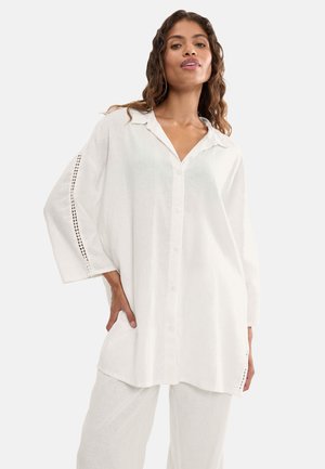 Kvinde iført en løs, lang hvid skjorte med knapper og udskæringsdetaljer på ærmerne, kombineret med matchende vidde bukser i hvid.