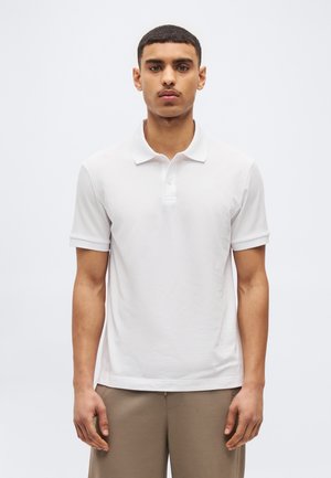 Muž s krátkymi tmavými vlasmi, oblečený v bielej krátkosleevej polokošeli a béžových nohaviciach, stojaci pred jednoduchým svetlým pozadím.