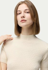 Ljus beige stickad tröja med ett strukturerat mönster, korta ärmar och hög krage. Slät tyg med en subtil glans.