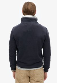 Marineblauer Pullover mit hohem Kragen und grauem Innenfutter. Mit einem Patch auf der Rückseite, gerippten Bündchen und Baumwollstoff. Lässiges Design.