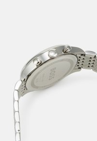 Montre en acier inoxydable avec un boîtier rond élégant, bracelet en maille argentée, trois poussoirs, et un dos gravé avec le logo "BOSS" et des détails de résistance à l'eau.