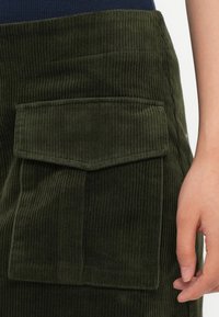 Detailní záběr na zelenou kordurovou sukni s velkým předním klopovým kapsou a částečný pohled na ruku osoby vedle ní.