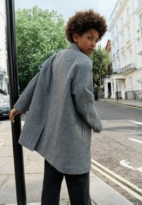 Oversized blejzer v šedém houndstooth vzoru s jediným rozventilovaním, uvolněnými klopami a dlouhými rukávy, spárovaný s tmavými kalhotami širokého střihu.