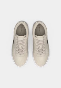 Ζευγάρι μπεζ αθλητικά παπούτσια Lacoste με κορδόνια, λευκές σόλες και ορατό λογότυπο στο εσωτερικό, απεικονιζόμενα από πάνω σε λευκό φόντο.