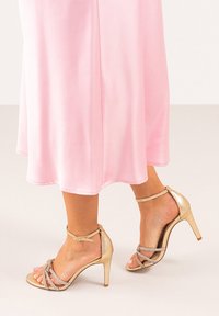 Sandalias doradas de tacón alto con correas cruzadas, una delicada correa en el tobillo y un acabado texturizado, llevadas con un vestido rosa fluido.