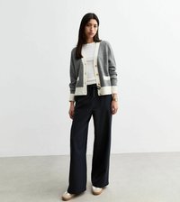 Grijze gebreide cardigan met witte accenten en knopen, gecombineerd met zwarte broek met brede streep en beige platform schoenen. Gladde textuur overal.