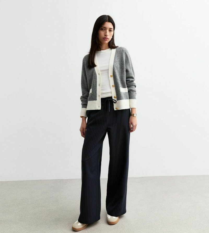 Grijze gebreide cardigan met witte accenten en knopen, gecombineerd met zwarte broek met brede streep en beige platform schoenen. Gladde textuur overal.