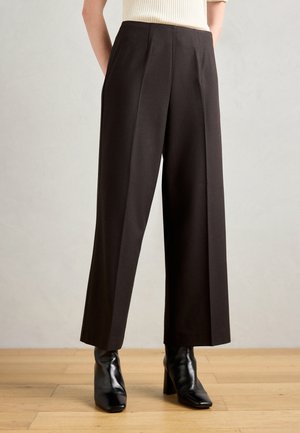 Un pantalon large noir associé à des bottines à talons noirs brillantes sur un fond de sol en bois clair.