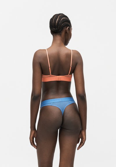 Γυναίκα με πλεξούδες, φαίνεται από πίσω, φορώντας ροδακινί σουτιέν και μπλε σχιστό εσώρουχο πάνω σε απλό λευκό φόντο.