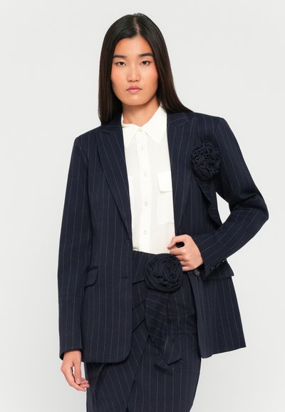 Kvinne iført en marineblå stripete blazer og skjørt med store stoffblomstdetaljer, kombinert med en hvit skjorte med knapper.