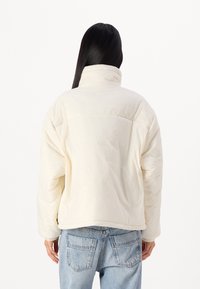 Pūdera jaka off-white krāsā ar augstu apkakli, kvadrātiem šūtu tekstūru un noliktām plecu daļām. Apvienota ar gaiši zilām džinsu biksēm.