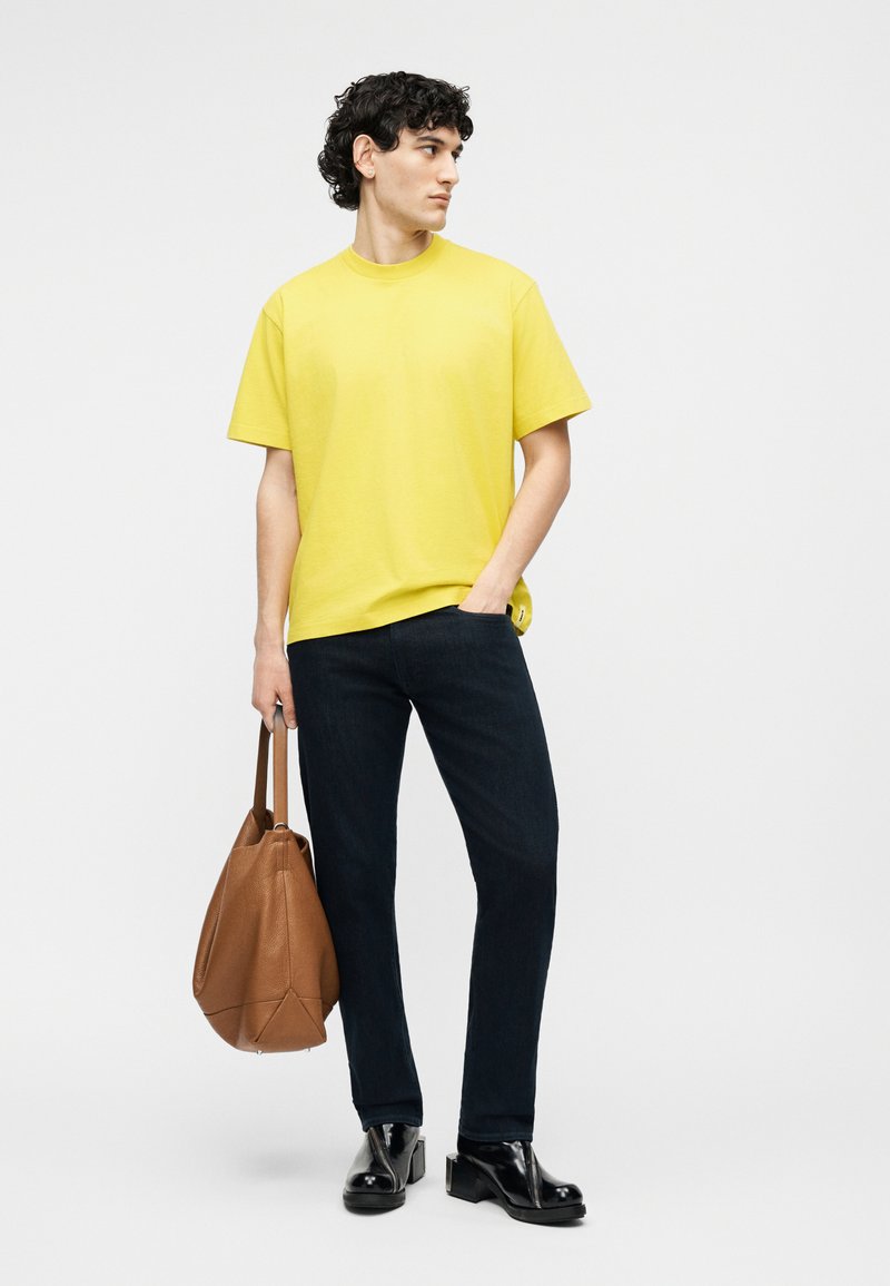 Uomo con maglietta gialla, jeans scuri e scarpe nere che tiene una borsa tote in pelle marrone, guardando di lato su sfondo semplice.