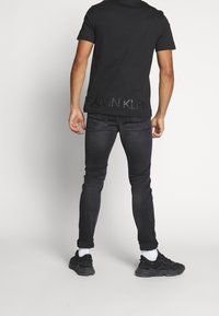 Černé bavlněné tričko s kulatým výstřihem a diskrétním potiskem loga Calvin Klein na zadní straně, zkombinované s tmavými slim-fit džíny a černými teniskami.