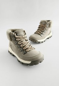 Beżowe buty trekkingowe z syntetycznej góry, kołnierzek wyłożony futrem, brązowe sznurowadła, metalowe oczka i solidna czarna gumowa podeszwa.