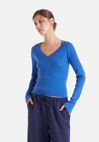 Suéter de punto acanalado azul con escote en V y mangas largas, combinado con pantalones oscuros de rayas. El suéter presenta un diseño ajustado y una textura suave.