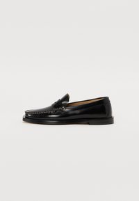Mocassins en cuir noir avec un bout arrondi, des coutures décoratives et une semelle plate. Texture lisse et design minimaliste.