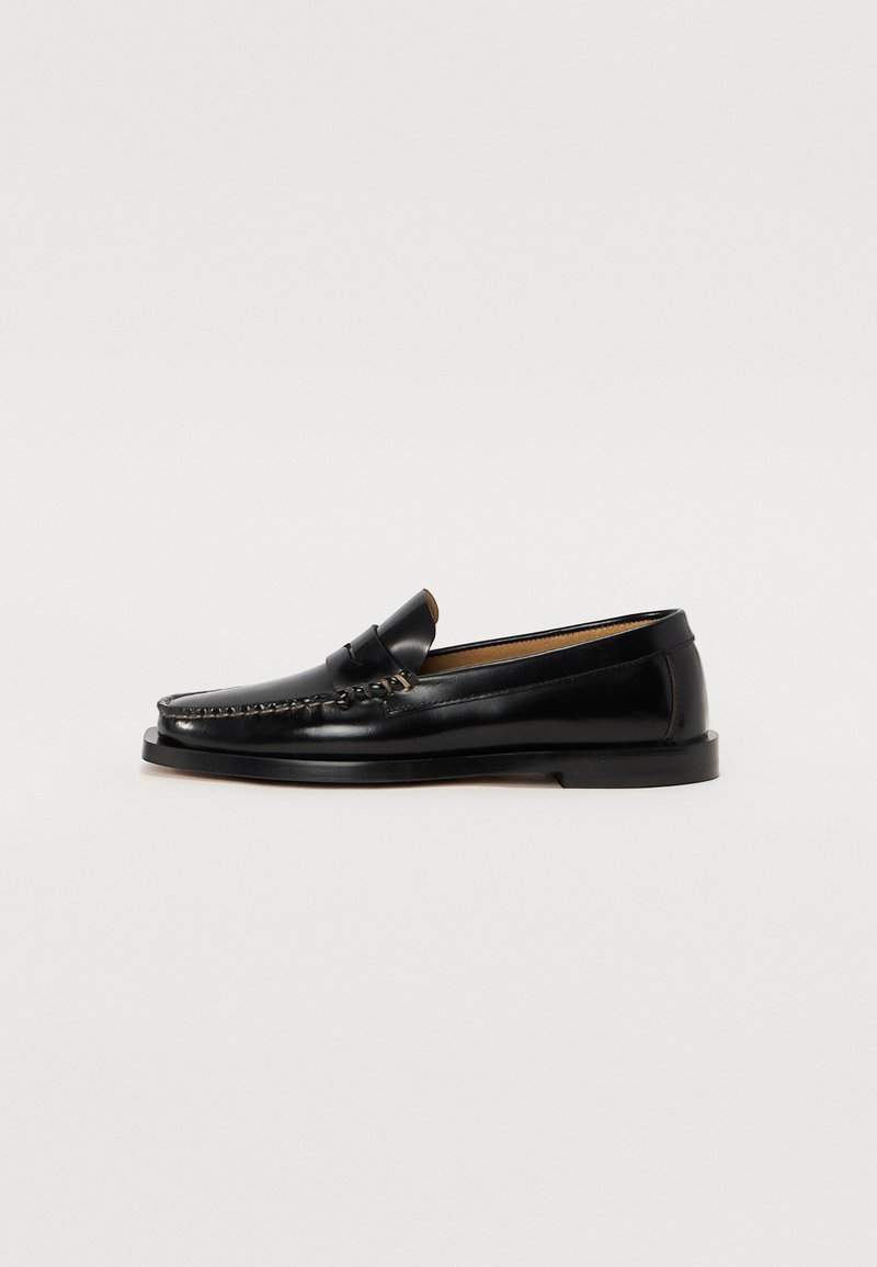 Mocassins en cuir noir avec un bout arrondi, des coutures décoratives et une semelle plate. Texture lisse et design minimaliste.