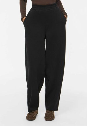 Personne portant un pantalon noir ample avec taille élastique et mains dans les poches, associé à un haut marron et des mocassins marron.