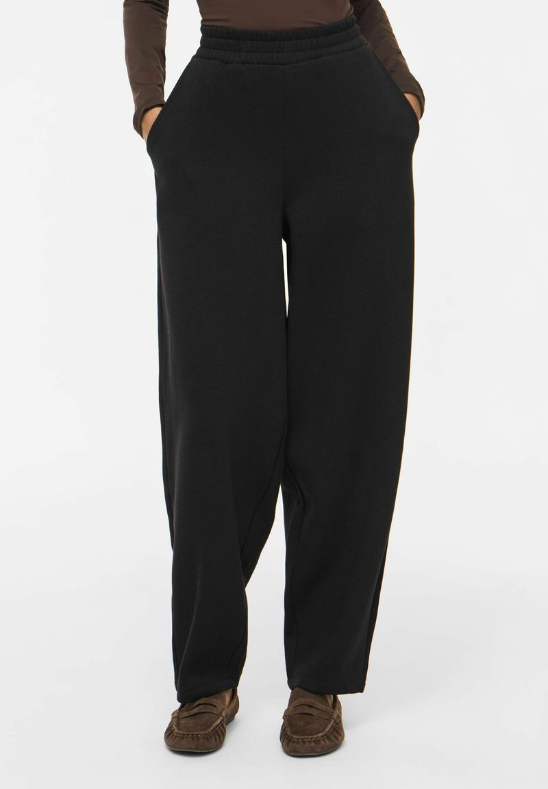 Personne portant un pantalon noir ample avec taille élastique et mains dans les poches, associé à un haut marron et des mocassins marron.
