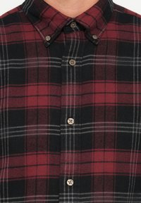 Camisa a cuadros en rojo y negro, con cuello abotonado, que lleva puesta una persona, mostrando solo el área del pecho y el cuello.