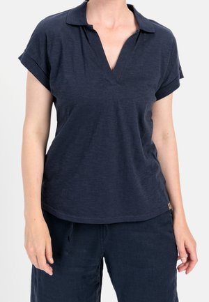 Persoon draagt een donkerblauw polo shirt met korte mouwen en een V-hals en donkerblauwe broek, staand tegen een effen achtergrond.