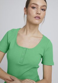 Top verde cu mânecă scurtă, cu model striat, decolteu rotund și deschidere cu trei nasturi. Materialul are o textură netedă și un design conformat.