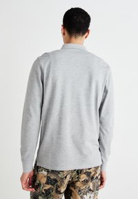 Graues langärmliges Poloshirt aus weichem Stoff, mit Kragen und seitlichen Schlitzen, kombiniert mit Hose im Camouflage-Muster.