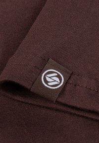 Brauner Stoff mit gerippter Textur. Verfügt über ein gewebtes Etikett in Dunkelbraun mit einem kreisförmigen weißen Logo. Nahaufnahme von Stoff- und Nähdetails.