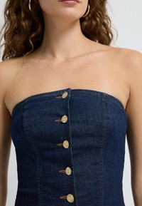Dżinsowy top bez ramiączek o dopasowanym kroju w ciemnoniebieskim kolorze, z sześcioma złotymi guzikami z przodu oraz widocznymi przeszyciami.
