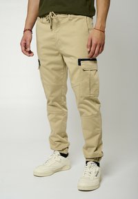 Pantalons cargo beiges avec des poignets élastiques, plusieurs poches et une taille à cordon, fabriqués à partir d'un tissu robuste au toucher lisse.