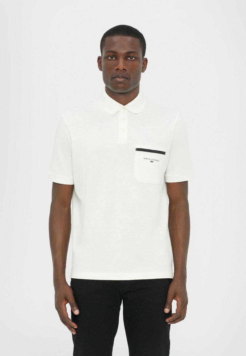 Hvit polo-shirt med krage, korte ermer og brystlomme med kontrasterende svart kant og logodetalj. Ensfarget stoffstruktur.