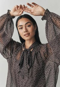 Pruunide polkadotidega pluus läbipaistvast kangast, köidetud kaelusega ja puffsiga pikkade varrukatega, millel on elastsed mansetid.