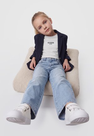 Une jeune fille est assise sur un fauteuil en maille couleur crème. Elle porte un blazer bleu marine à fines rayures, un tee-shirt blanc Guess, un jean large et des baskets blanches.