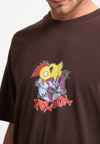 Brun bomulds-t-shirt med et farverigt grafisk motiv af en kylling på en globus indhyllet i pigtråd, med teksten "VOLCOM" nedenunder i rødt.