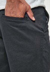 Pantalones gris oscuro con una textura suave, que cuentan con un bolsillo lateral y un ajuste casual. Primer plano muestra una mano metida en el bolsillo.