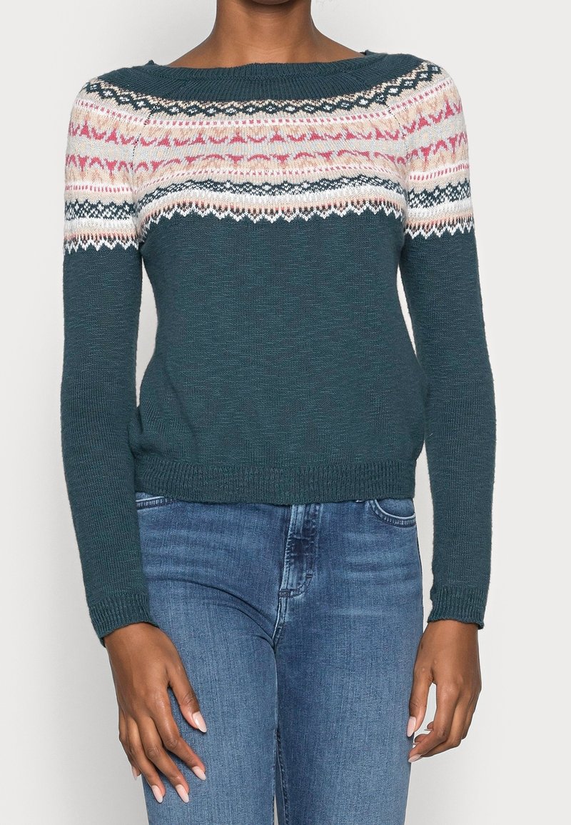 Pull tricot bleu sarcelle avec empiècement à motifs roses, blancs et noirs porté par une personne en jean bleu sur un fond uni.