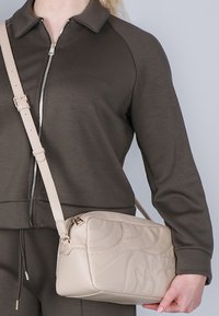 Beige crossover taske med kviltet tekstur og nittede detaljer. Har lynlås lukning og justerbar rem. Båret over et mørkegrønt outfit.