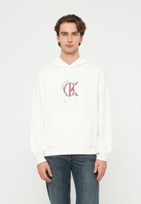 Tânăr cu păr ondulat, purtând un hanorac alb Calvin Klein și blugi albaștri, stând pe un fundal simplu și luminos.