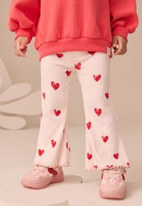 Svetloružové rebrované nohavice s červenými srdcovými vzormi, rozšíreným spodkom a volánikovým lemom, kombinované s ružovými teniskami s tmavším ružovým akcentom.