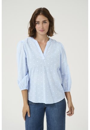 Helles blaues Baumwollbluse mit V-Ausschnitt, dreiviertellangen Puffärmel und einem strukturierten Punktemuster. Kombiniert mit dunkelblauen Jeans.